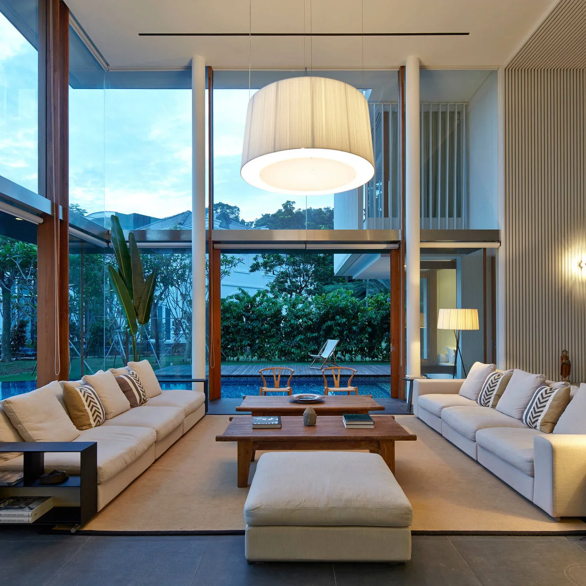 Two pale sofas face each other beneath a large drum pendant in a tall glass-walled living room with low wood tables.