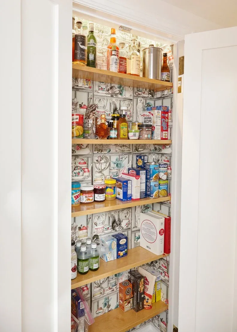 Small pantry with warm wood shelves, monochrome illustrated wallpaper and jars and packaged groceries