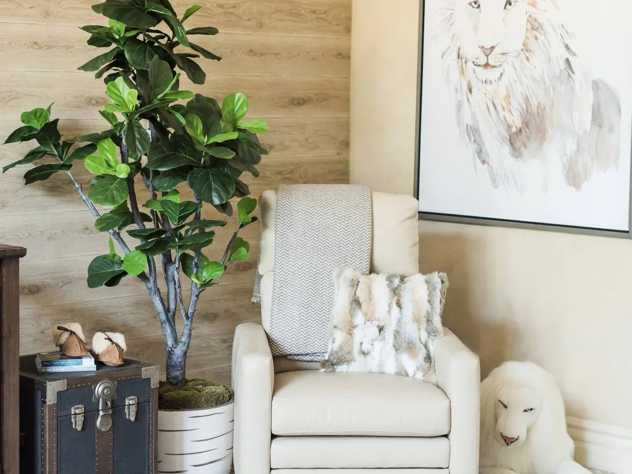 A cream nursery armchair sits beneath a large lion drawing beside a leafy plant and a plush white lion.