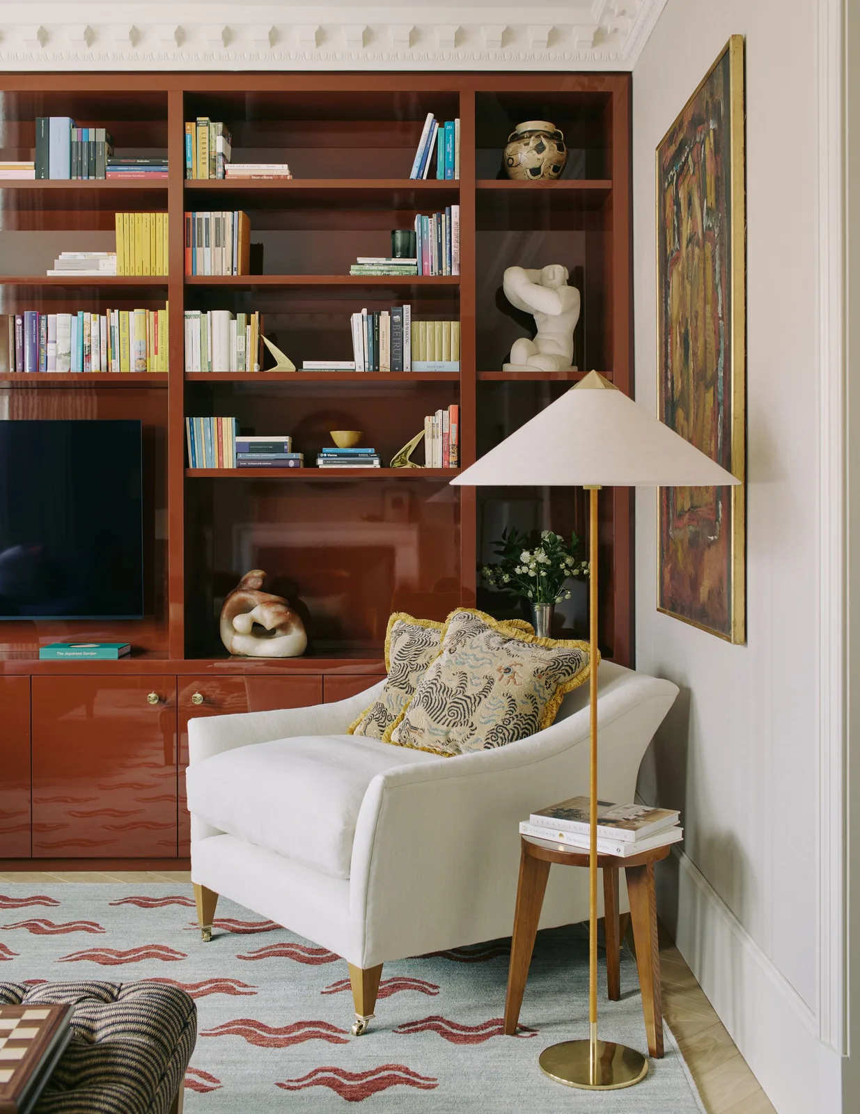 A television sits within glossy red-brown shelves and lower cabinets beside a pale armchair, patterned cushion and shaded floor lamp