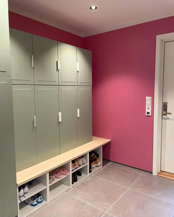 Muted green cabinet doors above a wood ledge and low white shoe cubbies beside a vivid pink entryway wall.