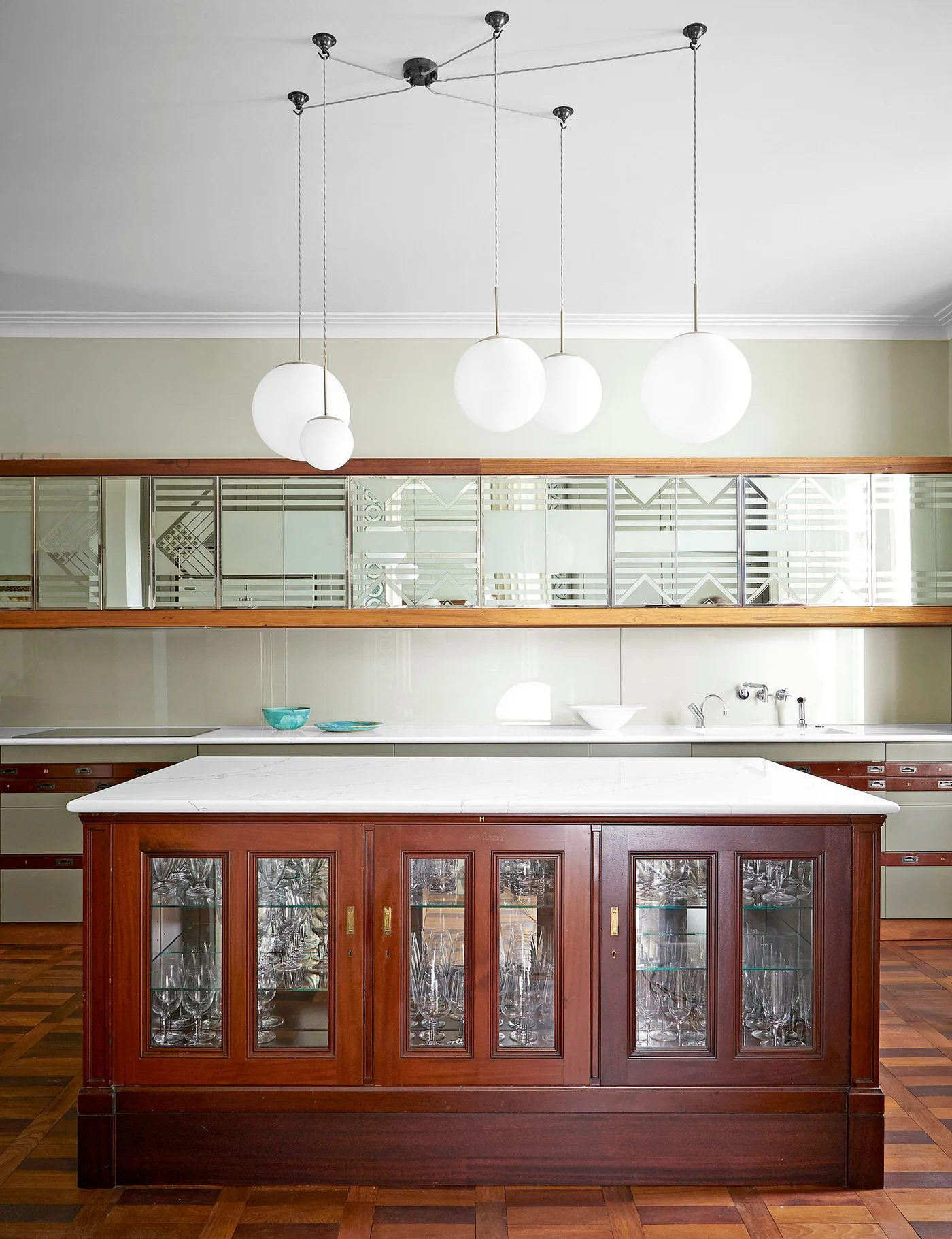 Kitchen with dark wooden glazed island, pale counter, long patterned mirrored wall and white globe pendants.