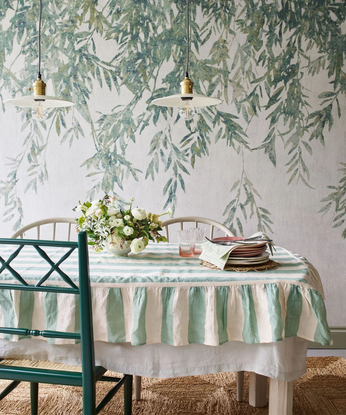 A soft green-striped cloth covers a small dining table beside mixed pale and green chairs below a leafy wall pattern