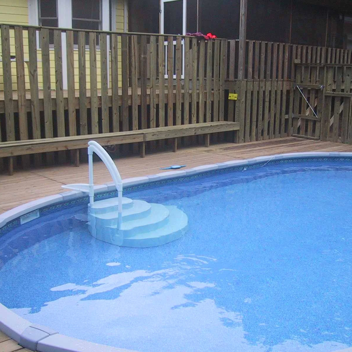 Broad white curved pool steps with a handrail beside a wood deck and a long narrow bench against the railing.