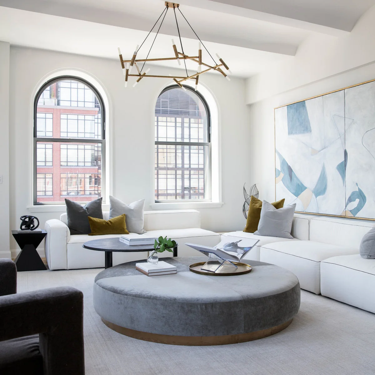 White sofas surround a large gray round ottoman with gray and olive cushions beneath pale abstract artwork.