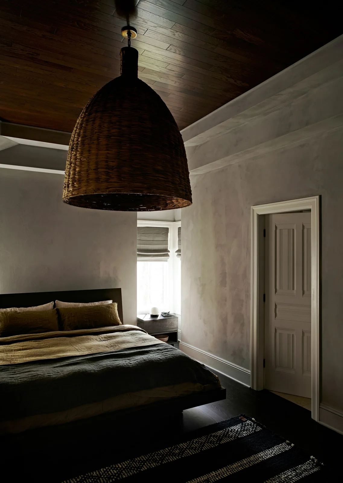 A bedroom has a dark wood ceiling, a broad woven pendant, pale mottled walls, a low dark bed and a small window beside a paneled door.