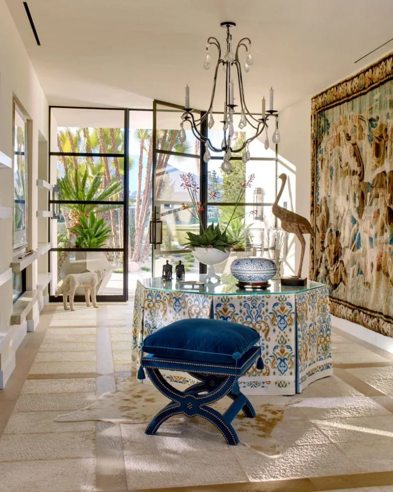 Dark curved candle chandelier with clear drops above patterned skirted table, blue crossed-leg stools and large wall tapestry in glazed entry