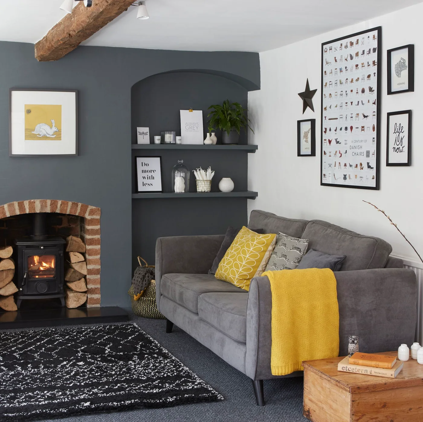 A gray sofa with yellow accents sits beside a brick-framed stove below an exposed wooden beam
