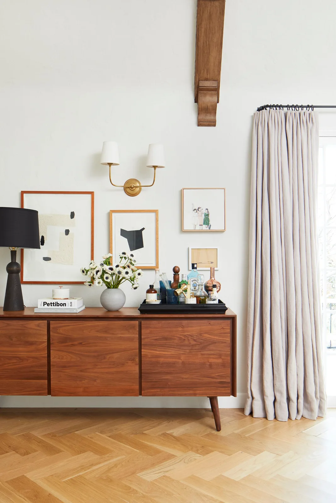 A wood sideboard stands below framed artworks with a black lamp, pale flower vase and drinks tray beside pale curtains
