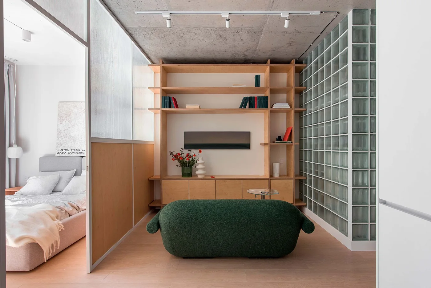 A small green sofa faces wood shelves and low storage beside a glass-block wall and glazed partition separating a bedroom