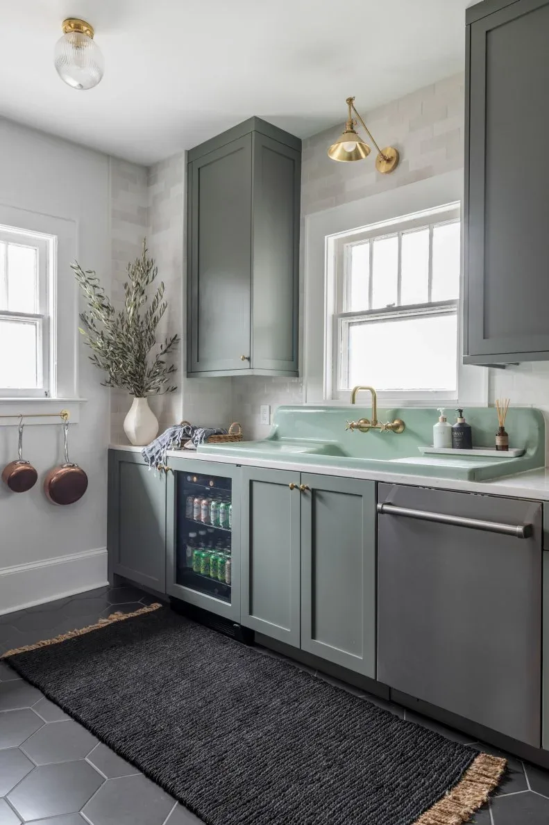 Gray-green kitchen cabinets with pale glossy backsplash tiles, a mint-colored sink with raised back and warm colored taps beneath a window.