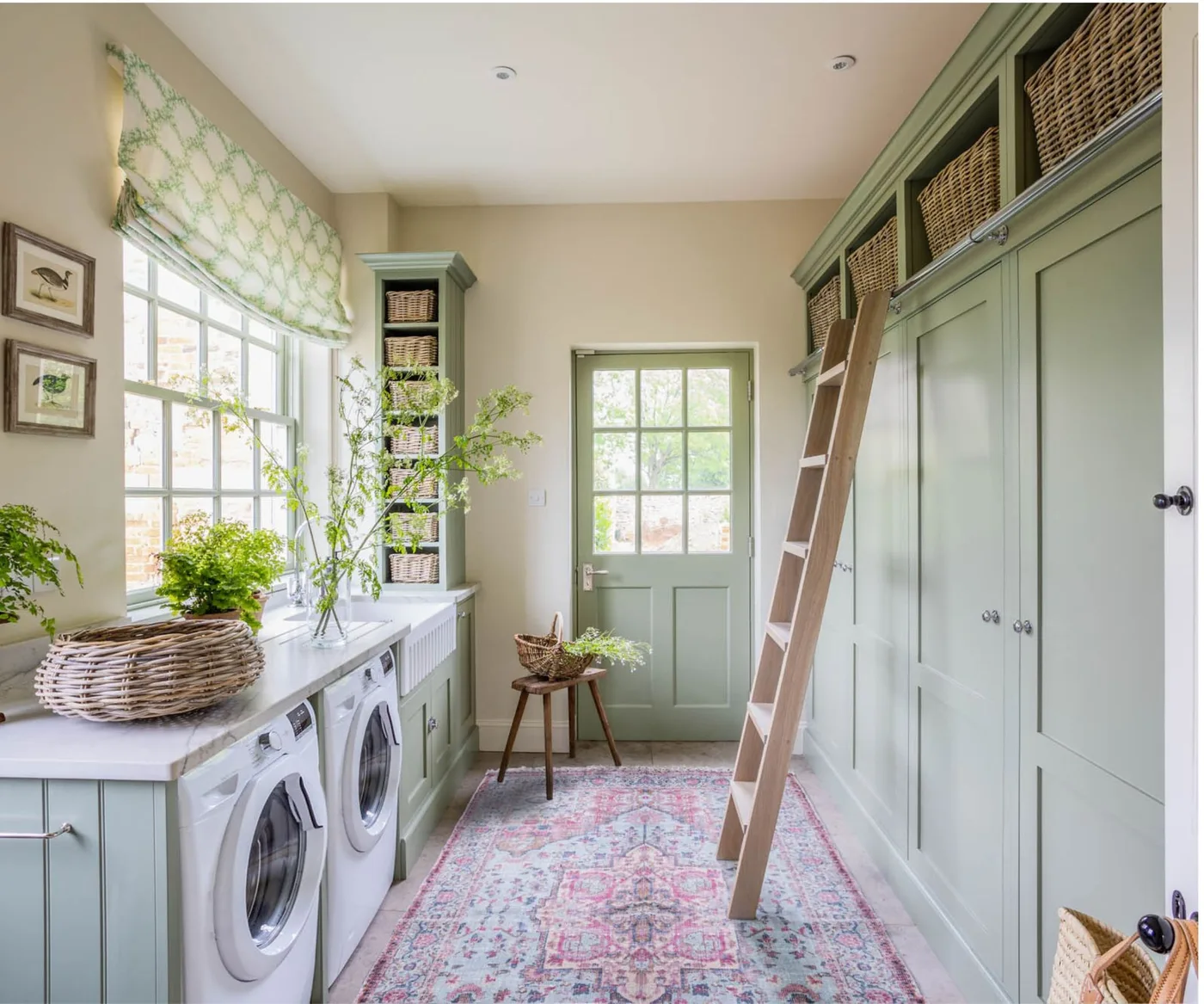 Sage green laundry cabinets with upper woven baskets, wood ladder, patterned rug and side-by-side appliances beneath a window