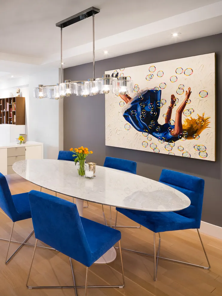 Blue upholstered chairs surround a slim pale oval table beneath a row of glass lights