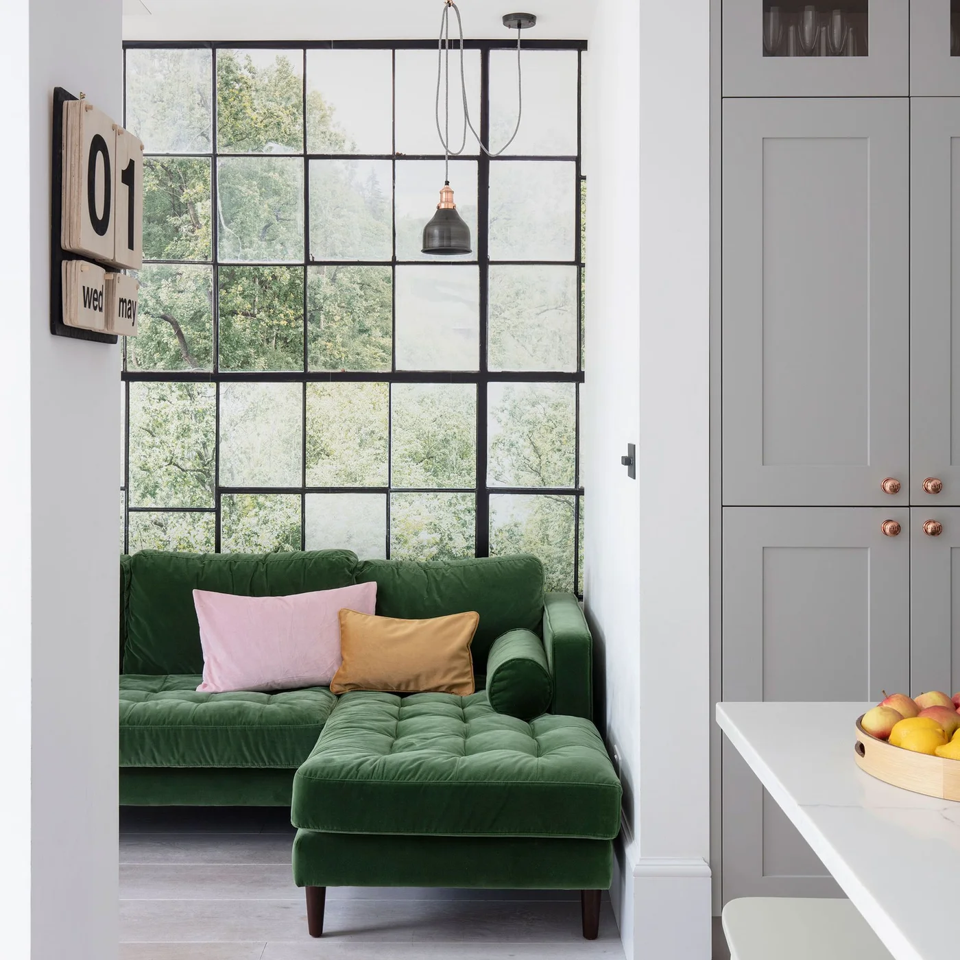 A woodland mural with a black window-like grid sits behind a green corner sofa beside gray kitchen storage