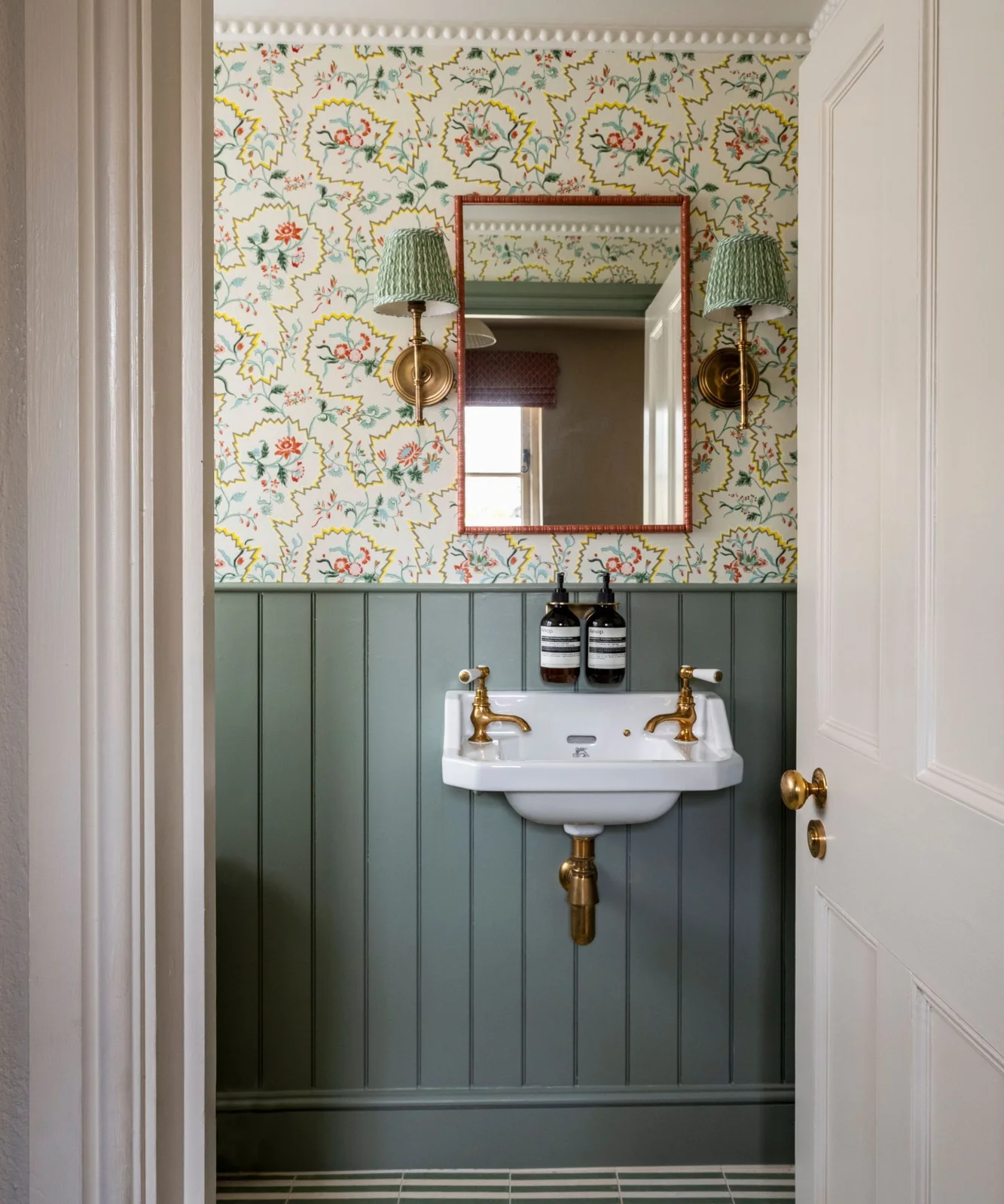 A tiny powder room has floral wallpaper above muted green vertical paneling, a small wall basin, red-edged mirror and green shaded wall lights.