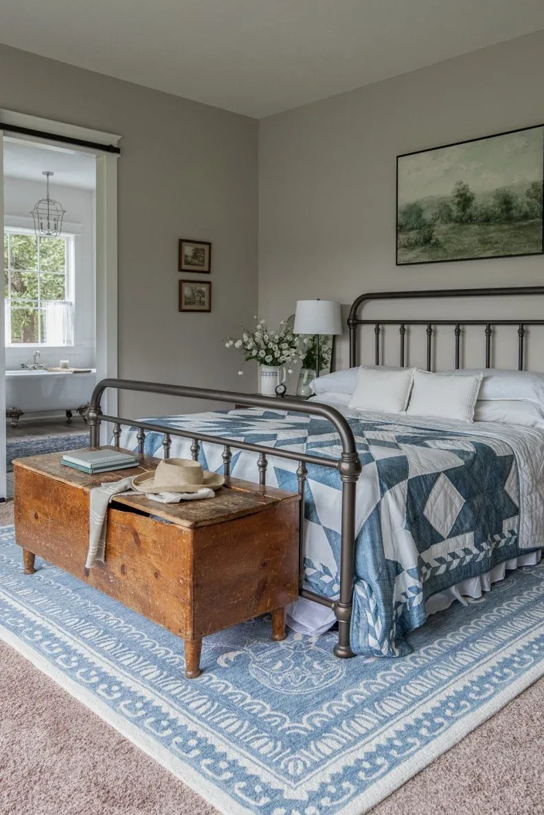 A metal bed with a blue patchwork quilt has a worn timber chest at its foot beside pale walls.