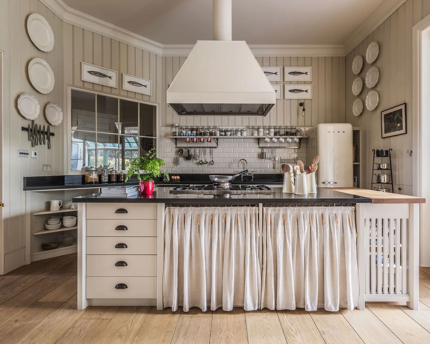 Kitchen with pale paneled walls, white central hood, curtained island front, drawers and wall-mounted plates.