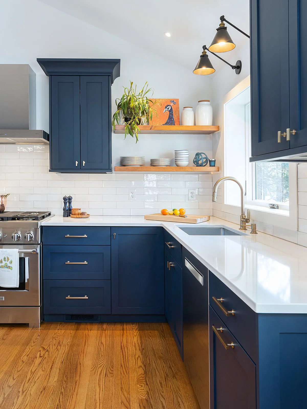 Blue lower and upper cabinets frame timber shelves with dishes against a pale tile wall beside the sink.
