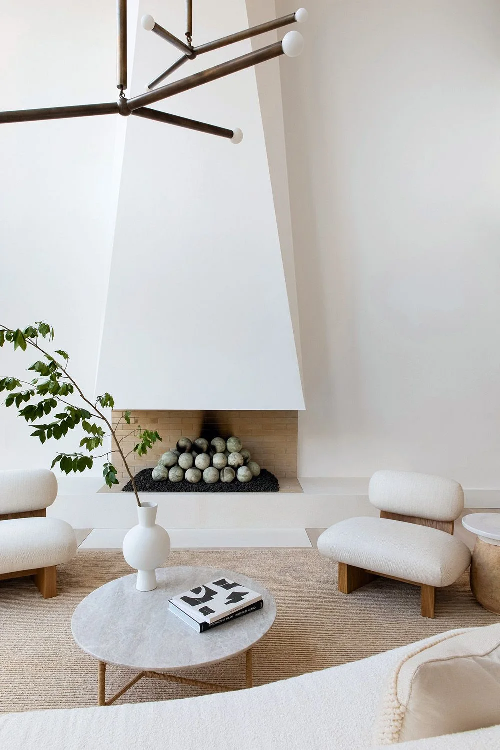 A tall tapered white fireplace form rises above a recessed opening with round decorative elements beside pale curved chairs and a small round table