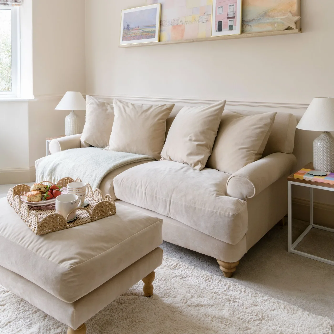 Pale warm-neutral sofa and matching footstool with a woven tray, simple lamps and a narrow picture ledge