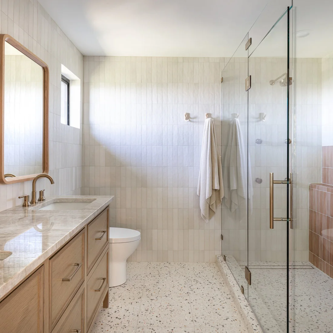 A pale wood vanity and rounded wood mirror face a glass shower with light vertical tiles, a pink bench and warm-metal fittings.