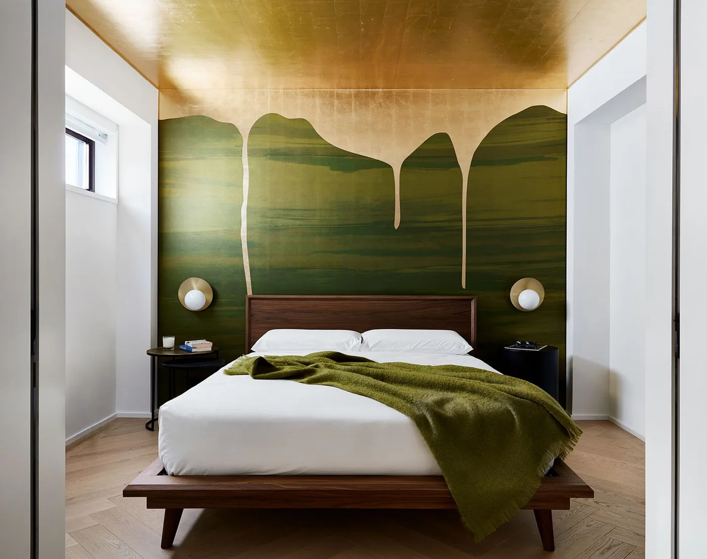 Modern bedroom with green and gold abstract wall decoration, a low wood bed, an olive blanket, globe wall lights and black bedside tables
