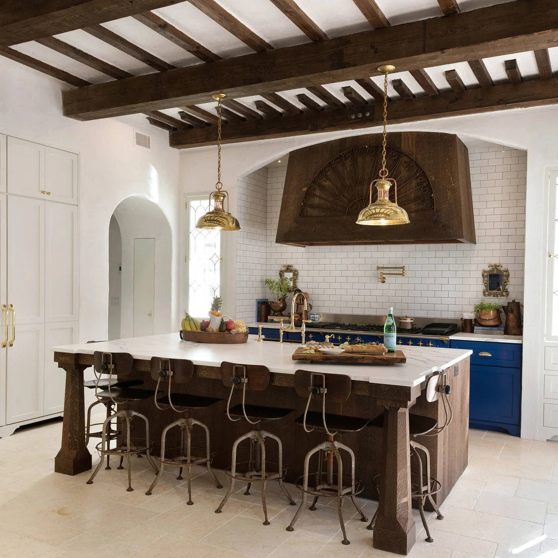 Dark ceiling beams and a broad decorative hood rise above a timber island, white cabinets and a blue range.