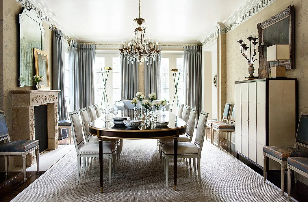 Oval dining table with pale chairs, a chandelier, a fireplace and tall gray curtains.