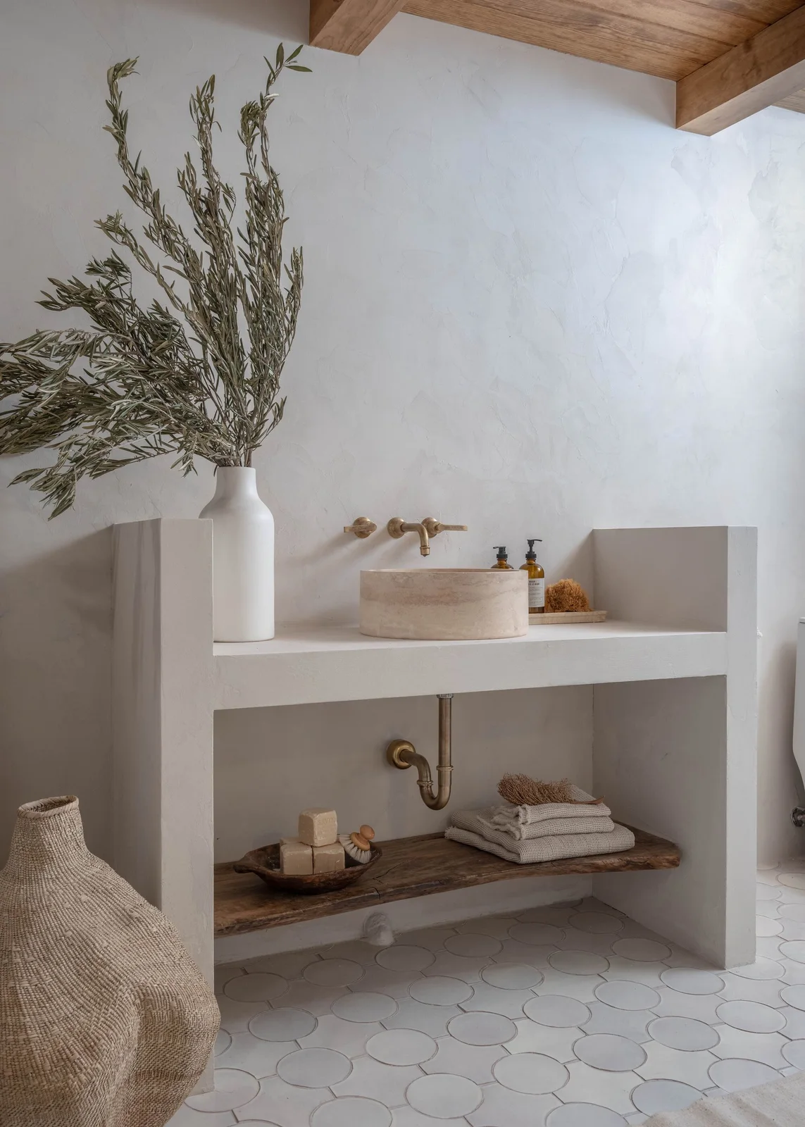 A pale cylindrical bowl sits on a white vanity with an open wood shelf, textured light walls and warm-metal wall taps.