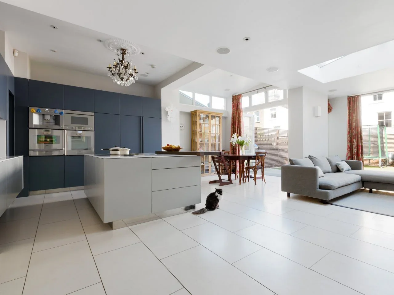 A dark-blue kitchen wall and pale island share a white tiled room with timber dining furniture and a gray sectional.