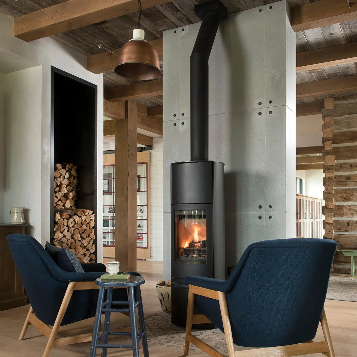 A tall curved-front black stove stands against a gray paneled wall in a timber-framed room with blue chairs and a separate tall log recess.