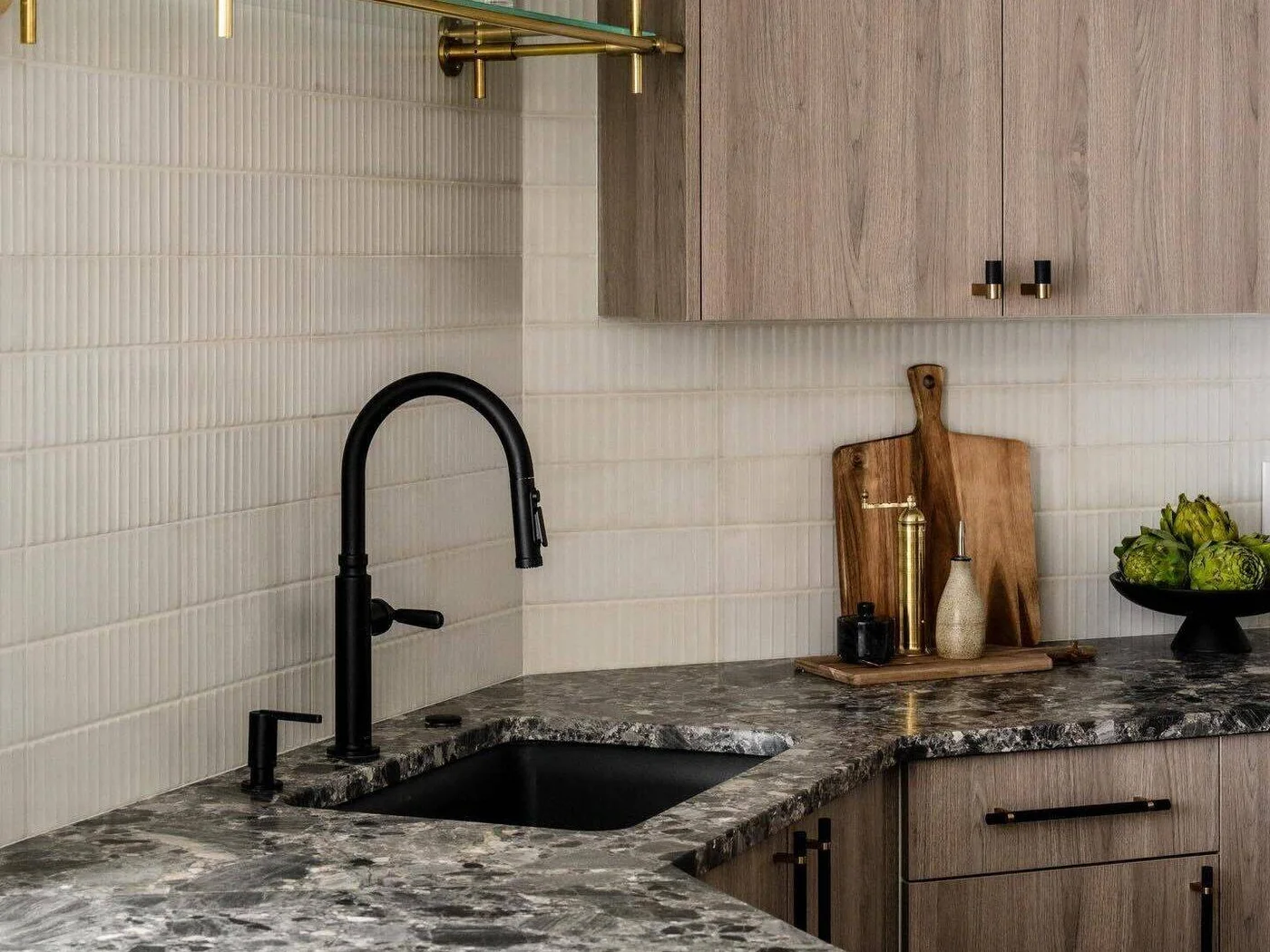 White fluted backsplash tiles sit above a dark veined counter beside pale timber cabinets and a black tap.