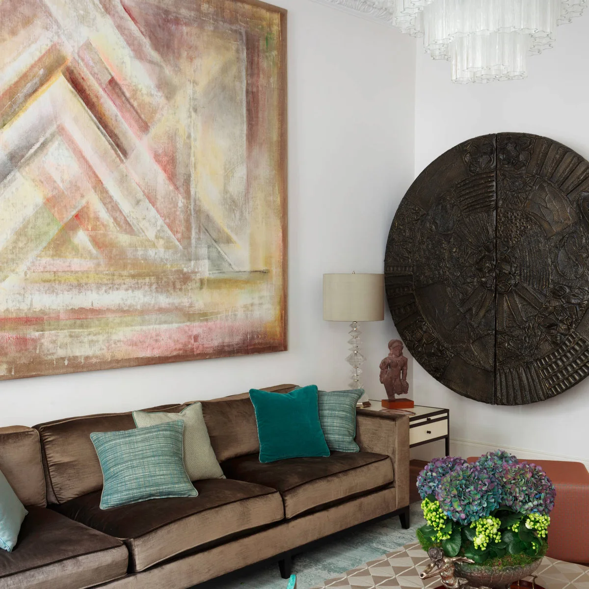 Brown fabric sofa with teal cushions below a large pale abstract painting beside a circular carved wall piece.