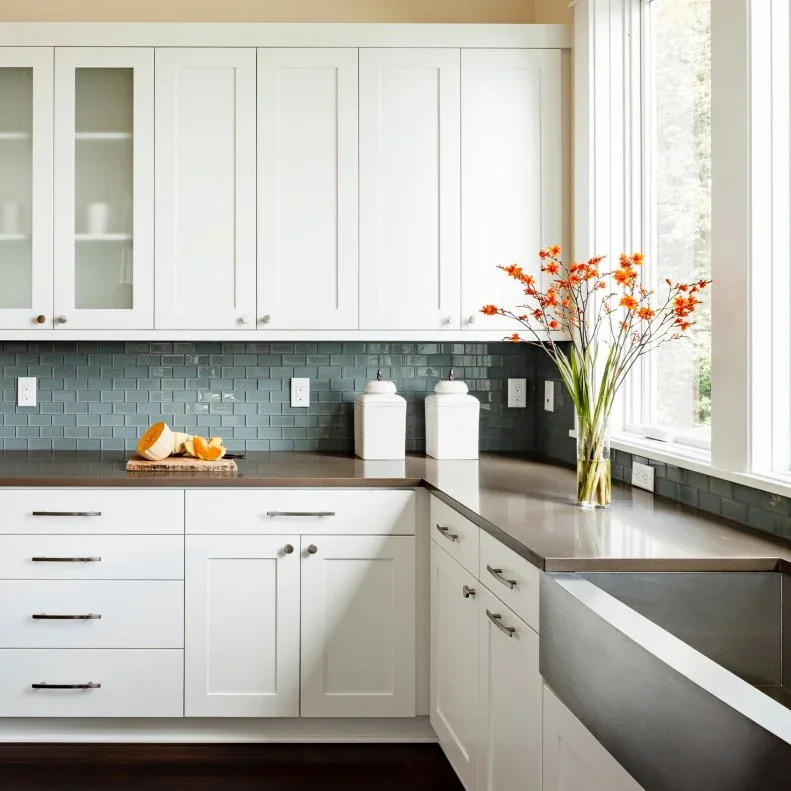 White framed cupboards and straight drawer handles stand above muted rectangular backsplash tiles and a darker counter beside a broad window