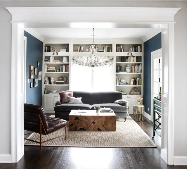 A charcoal sofa sits in front of a window framed by pale bookshelves, with a leather chair and timber coffee table nearby.