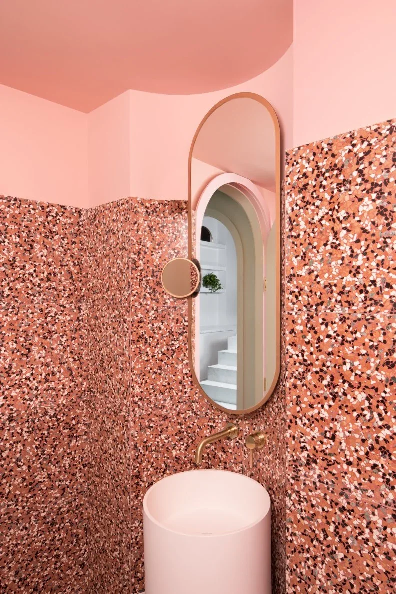 Pink terrazzo surrounds a pale pedestal basin below a tall oval mirror and plain pink walls.