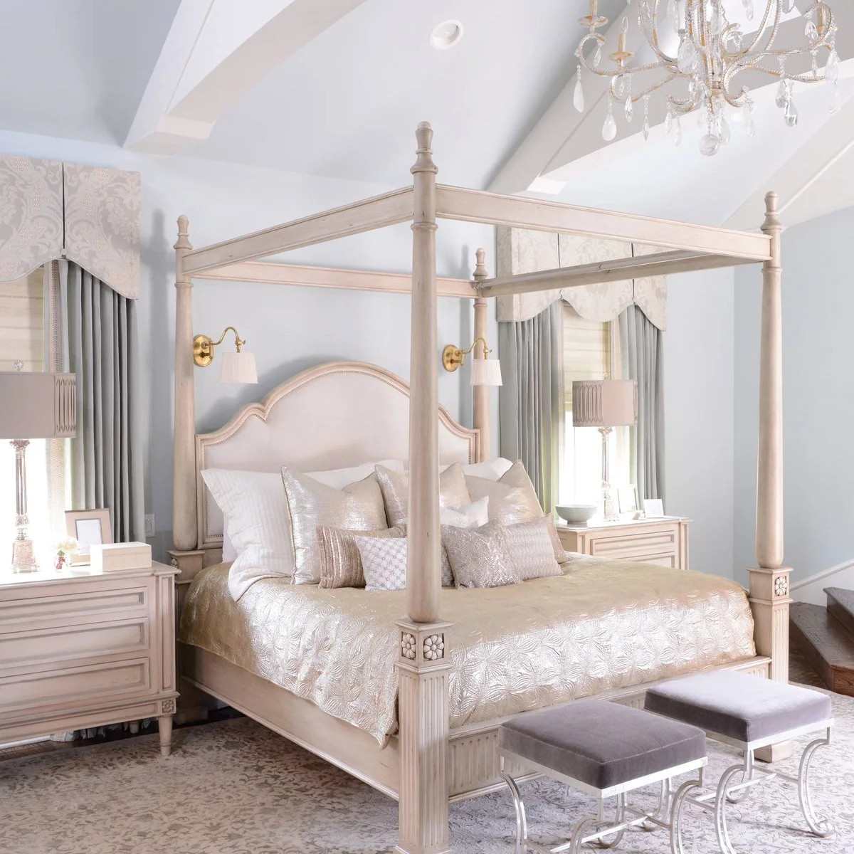 Pale four-poster bed with curved upholstered headboard, wall lamps and two small stools at the foot