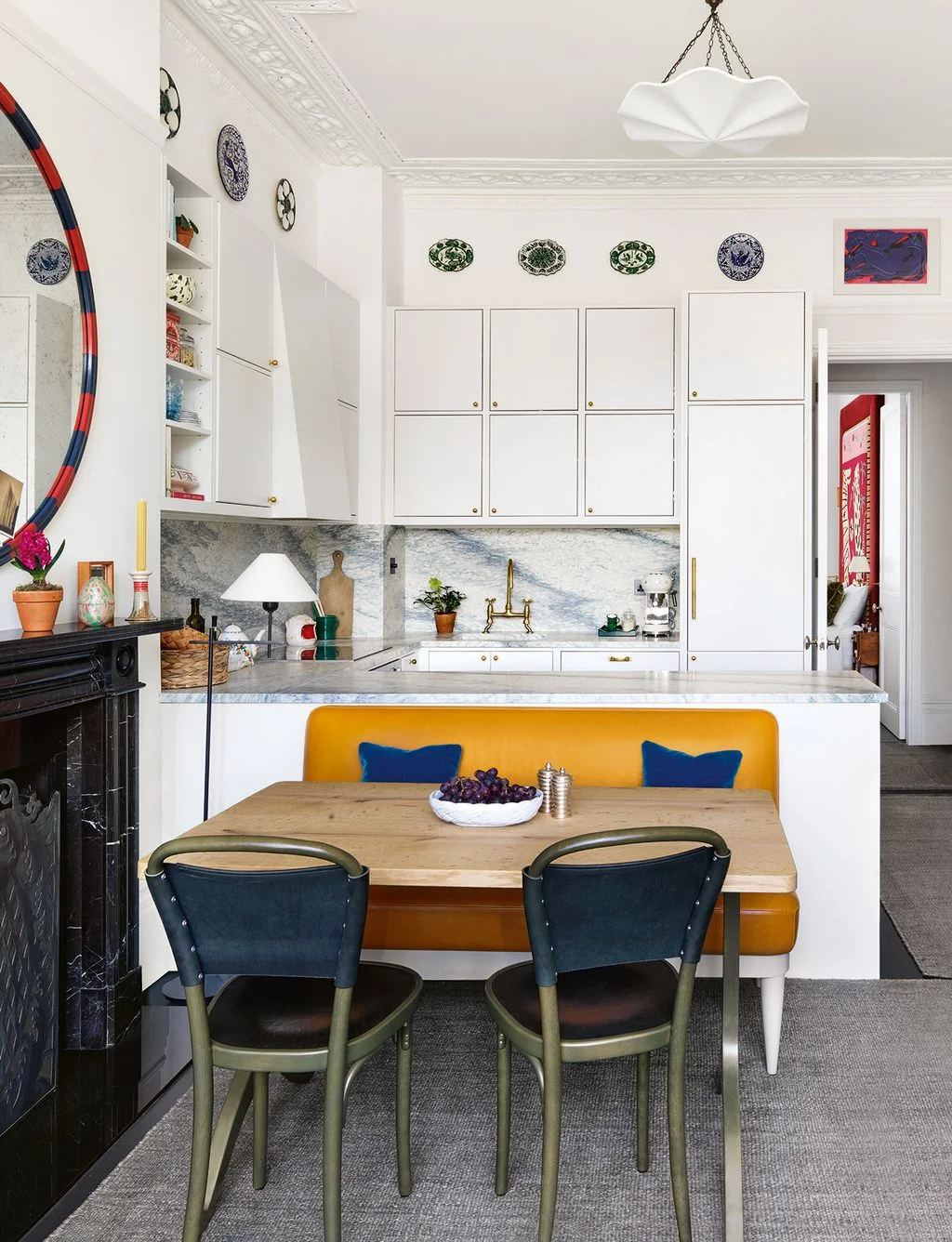 White kitchen cabinets sit beneath decorative plates and a sculptural pale pendant beside a yellow bench, blue cushions and a large colorful oval mirror