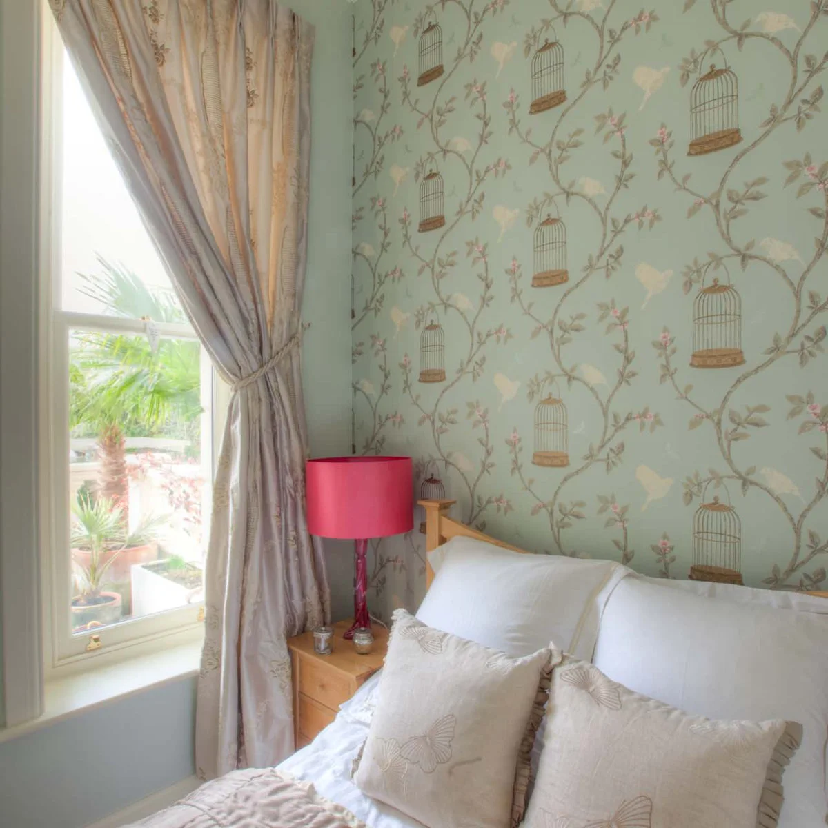 Pale green birdcage-and-branch wallpaper behind a wooden bed beside a window and a pink bedside lamp.