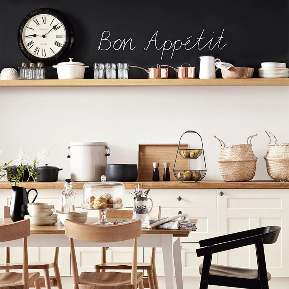 A kitchen dining corner has a dark upper wall with a clock and wood shelf above pale lower cupboards, warm counters and mixed light-and-dark chairs