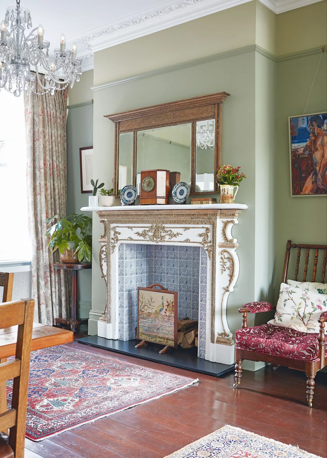 Pale ornate fireplace against green walls with wooden-framed mirror, chandelier, floral chair and patterned rug.