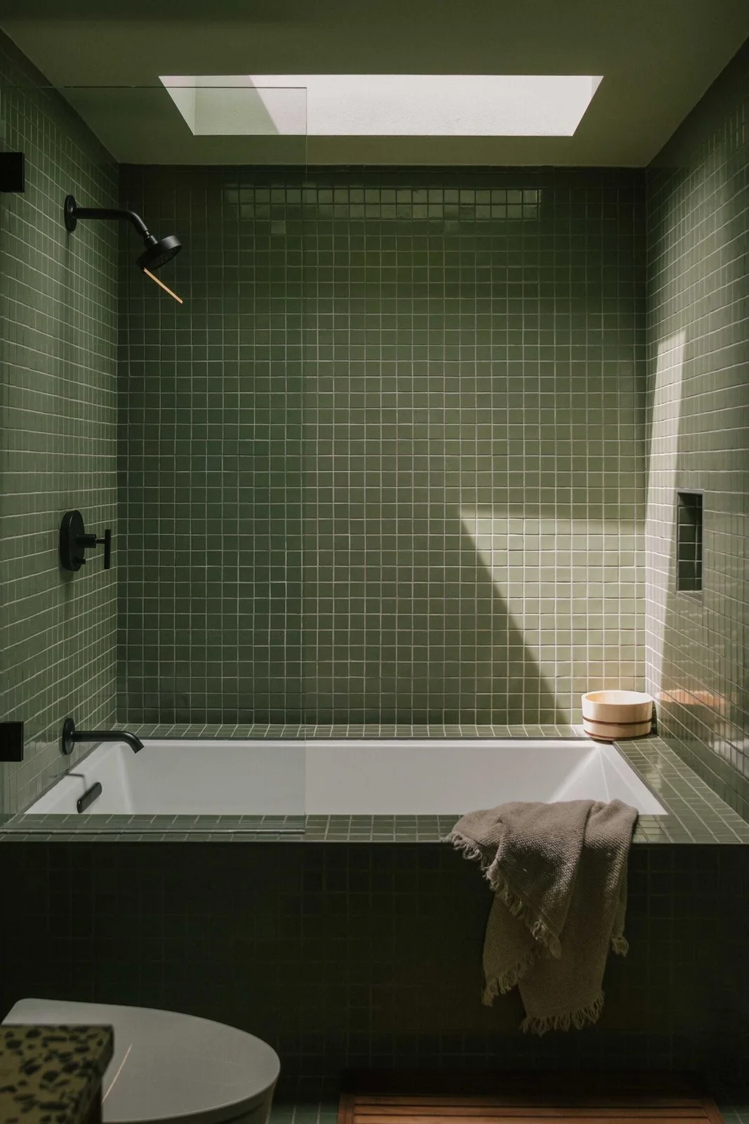 Small olive square tiles cover an inset bath surround and apron, with black fittings, a glass screen and a skylight above.