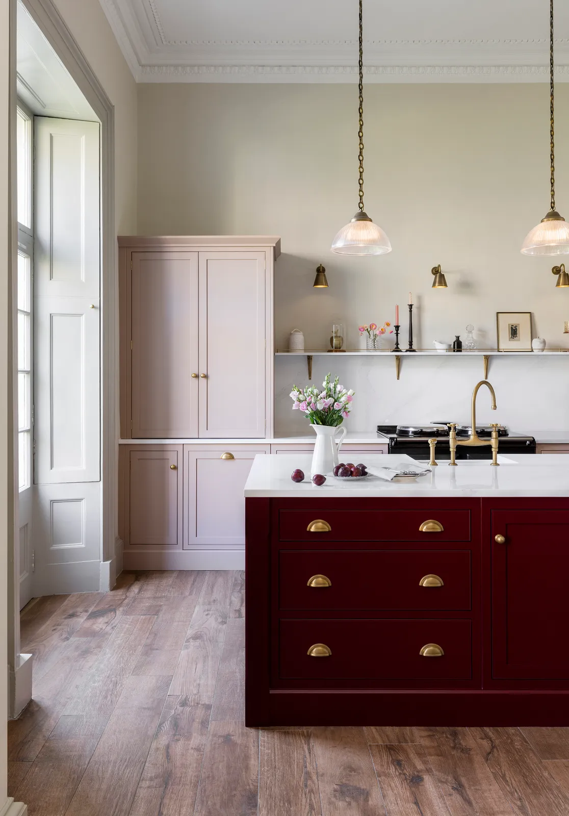 A burgundy island stands beside pale painted cabinets in a tall, bright kitchen