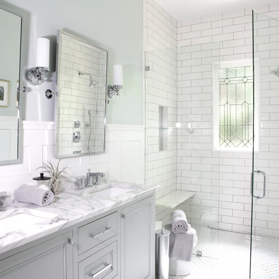 Small pale floor mosaics sit below white subway shower tiles, a gray vanity and a leaded window.