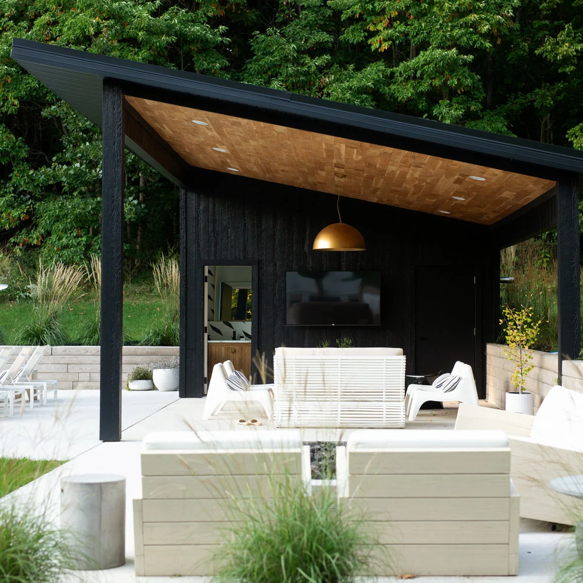 A dark freestanding single-slope pavilion has a warm wood-toned ceiling above white seating and a gold-colored pendant.