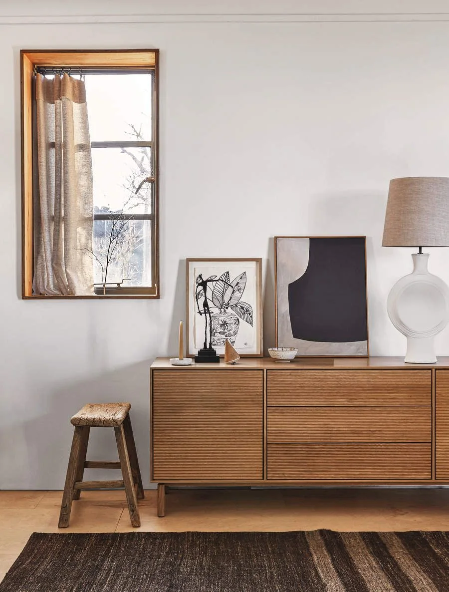 A low wood sideboard with finely ridged fronts stands below two leaning artworks and a white lamp beside a wood-framed window and small stool
