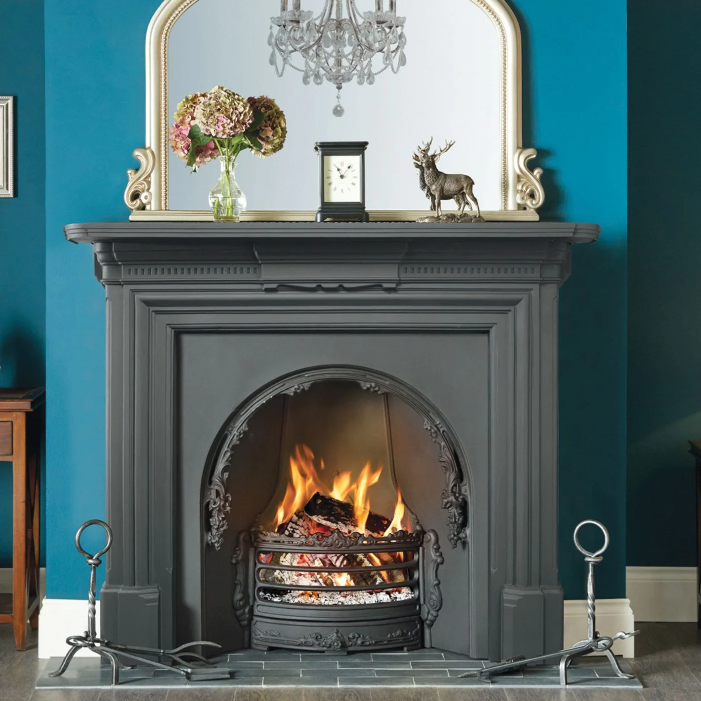 A dark gray molded fireplace with an arched opening stands against a teal wall beneath a large pale-framed mirror.
