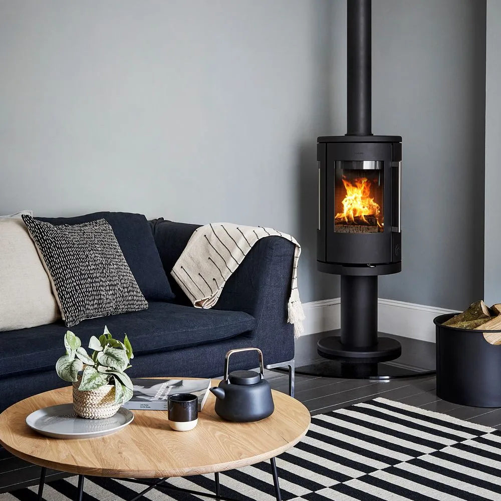 A black rounded stove stands on a dark hearth beside a blue sofa, with gray walls, a round wood coffee table, a black-and-white rug and a log container.