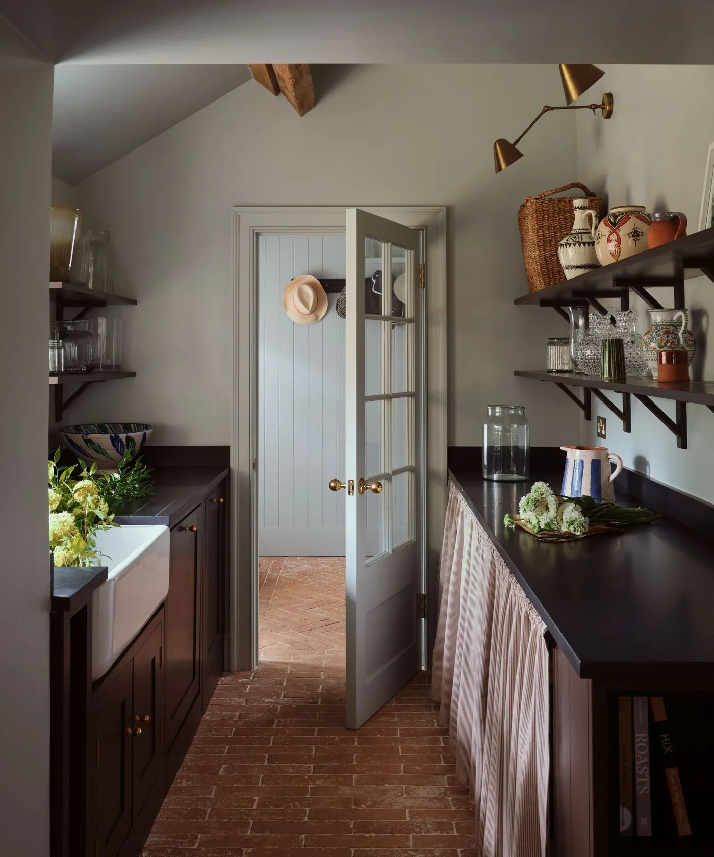 Narrow country pantry with dark shelves and counter, collected pitchers, pale lower curtain and white sink