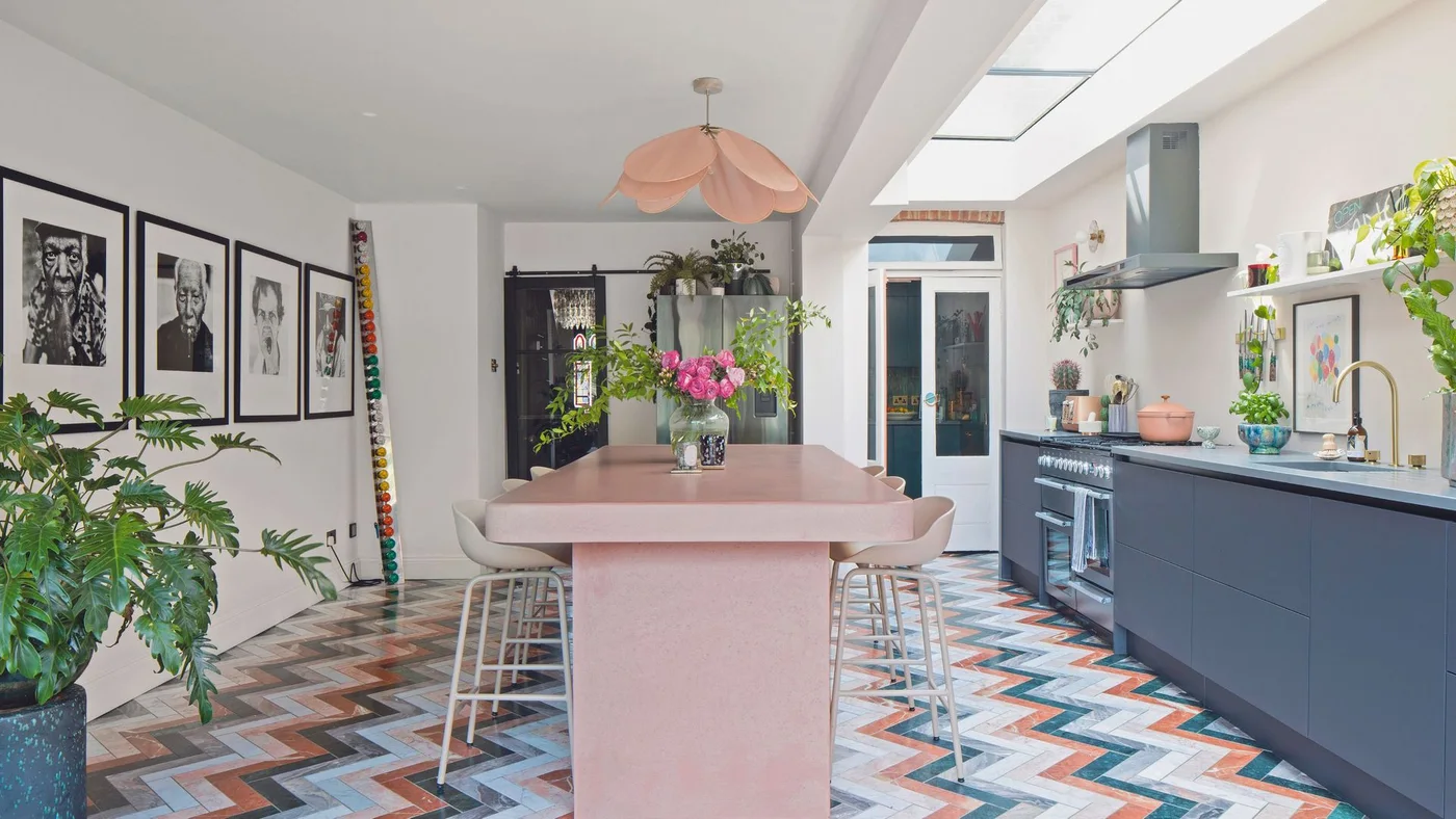 A pink island stands between dark blue cabinets and framed pictures above a multicolored chevron floor beneath a pale ceiling and pink pendant